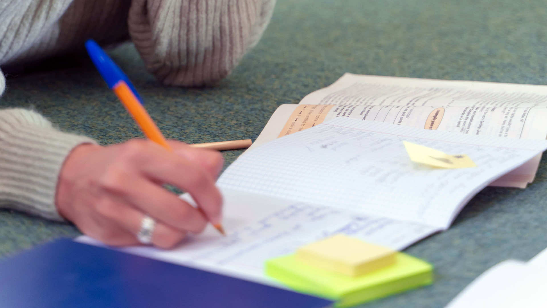 a woman's hand, writing in a notebook with a pen, with an open textbook nearby